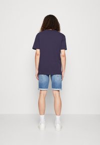 Marinblå bomullströja med korta ärmar, kombinerad med uppvikt denimshorts. Modellen bär vita strumpor och sneakers, sedd bakifrån.