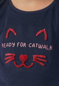 Marineblaues T-Shirt mit einem Katzenkopf-Design. Rote gestickte Akzente für Ohren, Schnurrhaare und ein Herz; pink glitzernder Text lautet "BEREIT FÜR DEN CATWALK."