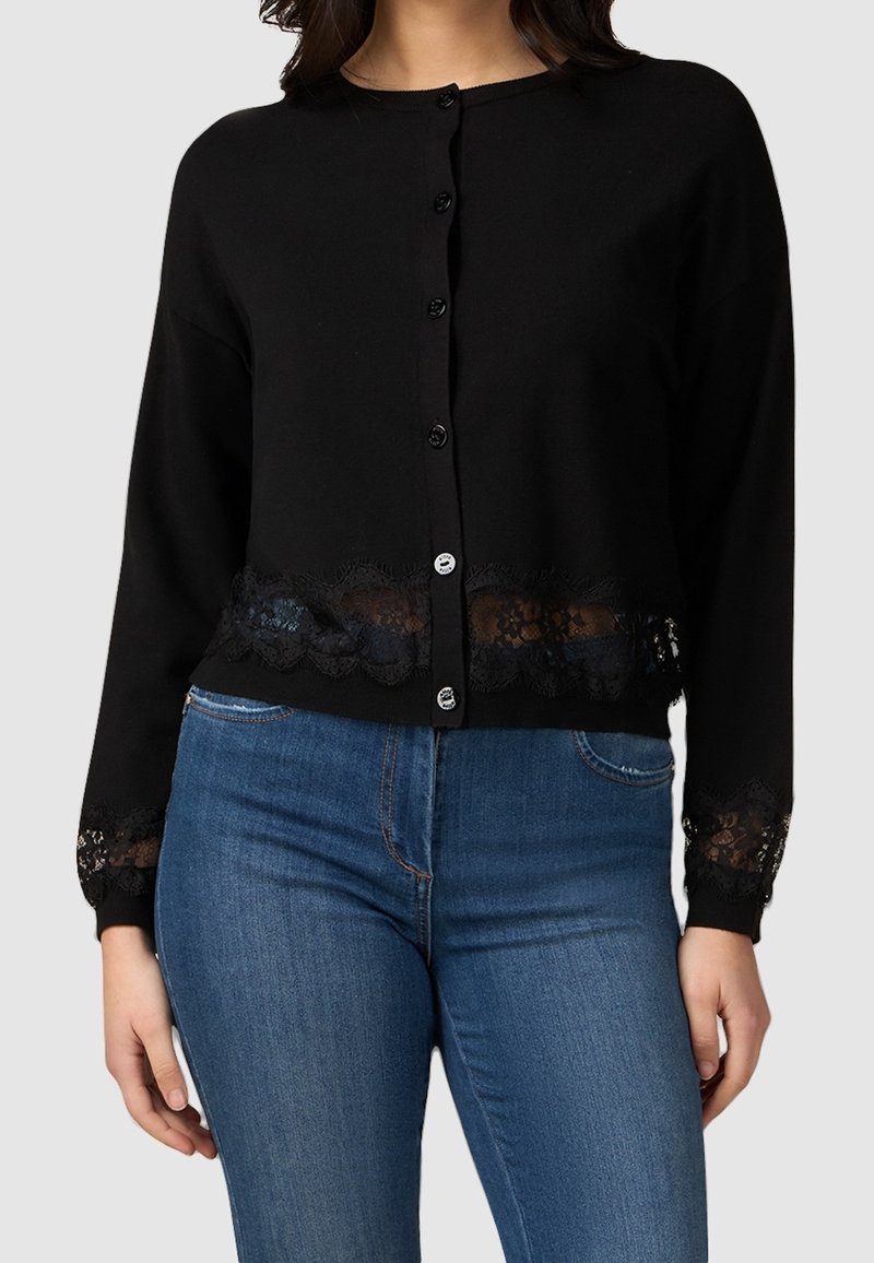 Black knit cardigan featuring a round neck, front buttons, and lace trim at the hem and cuffs. Paired with blue denim jeans.