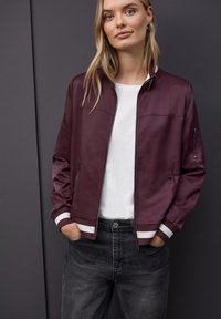 Veste bomber bordeaux avec des poignets et un col à rayures blanches, dotée d'une fermeture éclair à l'avant et de poches latérales, portée par-dessus une chemise blanche.