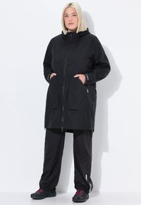 Donna con capelli biondi tagliati a caschetto che indossa una giacca nera con cappuccio e zip, con le mani in tasca, pantaloni neri e sneaker nere con lacci rosa.