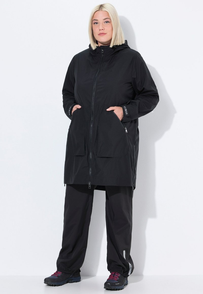 Donna con capelli biondi tagliati a caschetto che indossa una giacca nera con cappuccio e zip, con le mani in tasca, pantaloni neri e sneaker nere con lacci rosa.