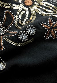 Tissu noir avec une broderie florale complexe ornée de sequins dorés, de perles et d'accents en fil brun, mettant en valeur la texture et le détail.