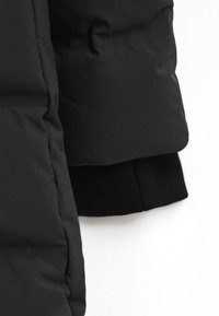 Svart puffjacka som visar slät tygtextur, quiltat mönster och ribbad stickad manschettdetalj vid ärmen.