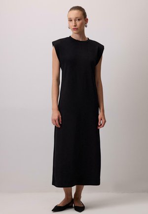 Žena stojí a má na sobě černé midi šaty bez rukávů se strukturovanými rameny a černé špičaté baleríny na jednobarevném pozadí.