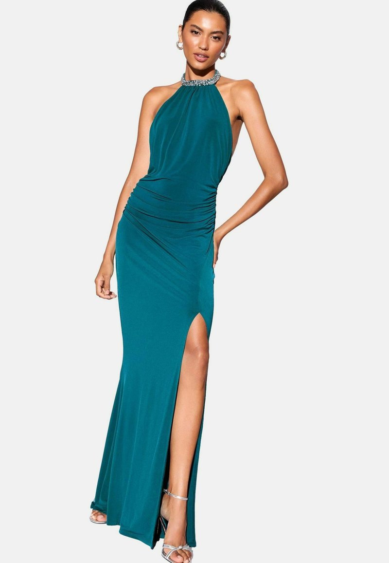 Frau in einem ärmellosen, türkisfarbenen Abendkleid mit einem perlenverzierten Halsband und einem hohen Schlitzen bis zum Oberschenkel, kombiniert mit silbernen High-Heels.