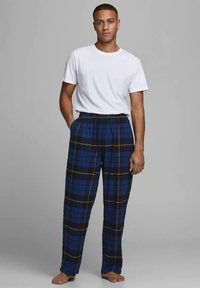 Ung mann som står barbeint iført en enkel hvit t-skjorte og blå rutete pysjamasbukser med hendene i lommene mot en grå bakgrunn.