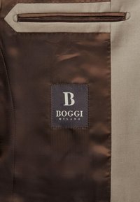 Brūnas un bēšas krāsas audums ar BOGGI Milano logo plāksnīti. Iekļauj kabatu ar kontrastējošu apdari. Gluda tekstūra un pielāgots dizains.
