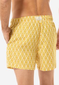 Pantaloni de baie galbeni cu un model în romb alb, cu talie elastică și buzunare laterale. Materialul pare ușor și neted.