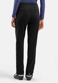 Personne portant un pantalon de sport noir et des chaussures de sport noires, se tenant de dos face au spectateur sur un fond blanc.