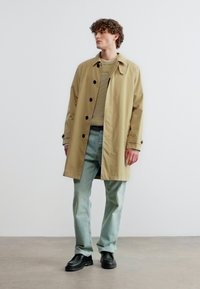 Homme portant un trench-coat beige, un pantalon vert clair, un pull à rayures et des chaussures noires, debout contre un mur uni.