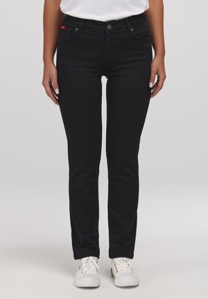 Kobieta ubrana w czarne przylegające dżinsy i białe trampki, stojąca na tle jednolitego szarego tła.