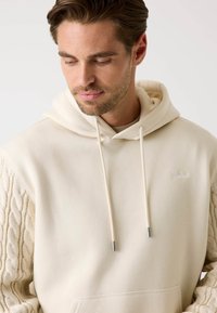 Sudadera beige con mangas de punto trenzado, capucha con cordón y bolsillo delantero. Presenta un logotipo sutil en el pecho y una textura de tela suave.