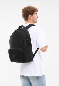 Svart ryggsäck i tyg med rundad design, framfack med dragkedja och justerbara vadderade axelremmar. Bärs på ryggen.