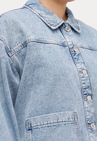 Lichtblauwe spijkerjack met een puntkraag, knopen aan de voorkant en een borstzak. Heeft een losse pasvorm en zichtbare stiksel details.