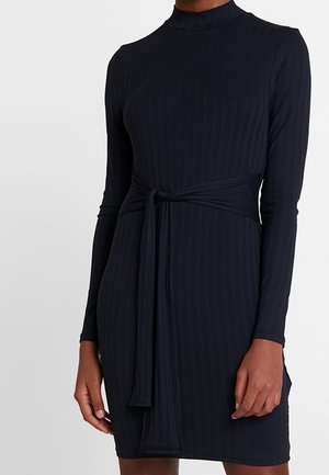 Frau trägt ein langärmeliges, hochgeschlossenes schwarzes Kleid mit vertikaler Rippenstruktur und einem gebundenen Gürtel in der Taille.