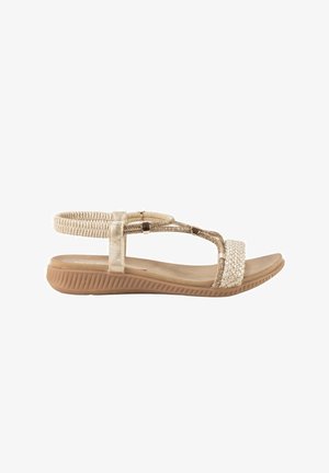 Sandalia plana beige con correa elástica en el tobillo, tira frontal trenzada y suela marrón texturizada diseñada para uso casual.