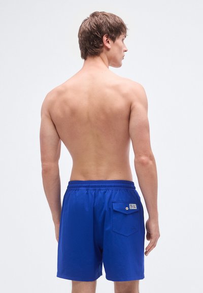 Μπλε μαγιό βερμούδα με ελαστική μέση, που έχει πίσω τσέπη και λεπτομέρεια με ταμπελάκι, εμφανιζόμενο σε άτομο που φαίνεται από πίσω.