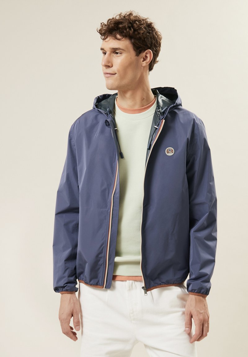 Navy wasserfeste Jacke mit Kapuze, Reißverschluss und elastischen Bündchen. Mit einem Logo auf der Brust und getragen über einem hellgrünen Hemd.