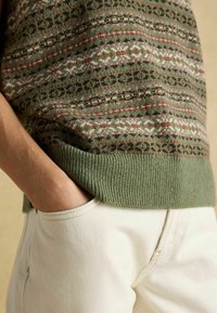 Isik, kes kannab roheline muster kootud sviit, käsi selja taskus heleda beeži pükste püsib neutraalses taustas.