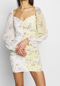 Vestido mini floral con escote corazón, que presenta un lado texturizado blanco y un patrón floral amarillo, mangas abullonadas y detalles fruncidos.