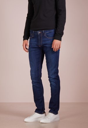 Homme portant un jean slim bleu foncé, des baskets blanches et un t-shirt à manches longues noir debout sur un sol et un fond beige.