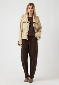 Femme debout portant une veste beige à volants, un pull marron, un pantalon marron ample et des bottines en daim marron, faisant face à l'avant sur un fond uni.