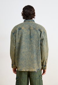 Giacca di denim in verde-blu sbiadito con colletto abbottonato, ampia spalla e maniche risvoltabili; abbinata a pantaloni cargo color oliva.