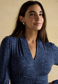 Nais, kellel on pikad tumedad juuksed, kannab kuldseid rõngaste kõrvarõngaid ja sinist mustriga V-kaelusega kleiti, seistes heledal beežil taustal.