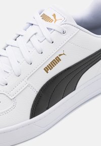 Baltas sintetinis odos sportbačių modelis su juodu juostos akcentu, auksiniu Puma logotipu ir baltais raišteliais. Pado spalva balta, su tekstūrizuota apdaila.