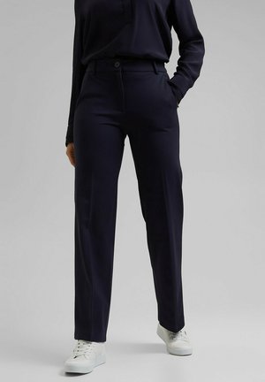 Personne portant un pantalon droit bleu marine foncé, un haut foncé assorti et des baskets blanches, debout devant un fond clair uni.