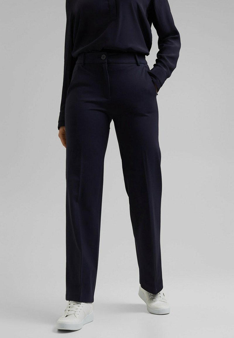 Personne portant un pantalon droit bleu marine foncé, un haut foncé assorti et des baskets blanches, debout devant un fond clair uni.
