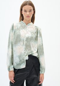 Blusa verde chiaro con righe verticali e un motivo floreale, dotata di maniche lunghe, colletto alto e chiusura con bottoni davanti.