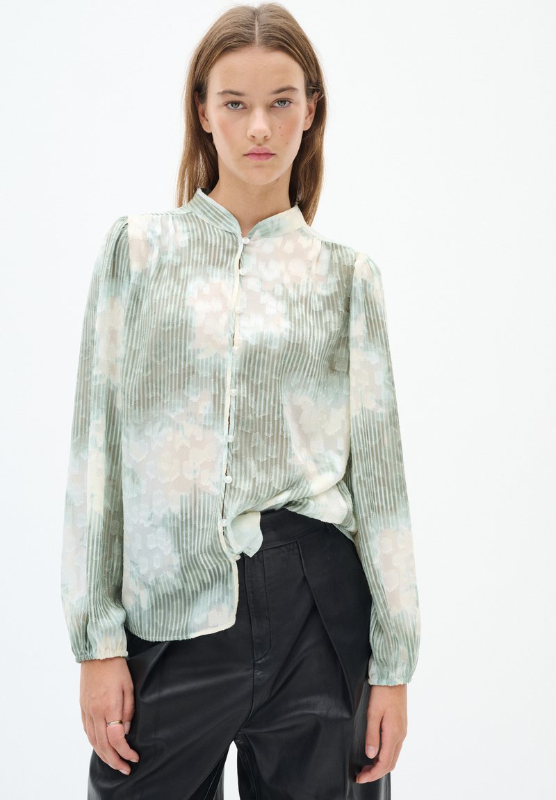 Blusa verde chiaro con righe verticali e un motivo floreale, dotata di maniche lunghe, colletto alto e chiusura con bottoni davanti.