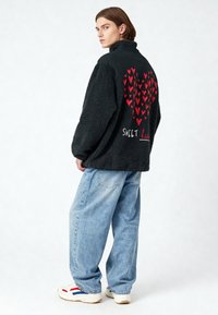 Jeune homme portant une veste polaire noire avec un groupe de cœurs rouges et le texte "SWEET heart", un jean large bleu clair et des baskets blanches, debout de profil.