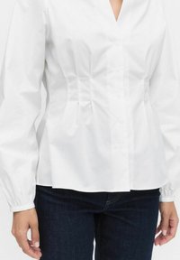 Vit skjorta med knappar och v-ringad halsringning samt veckad midja. Har långa, elastiska ärmar och mjukt bomullstyg. Kombineras med mörka jeans.