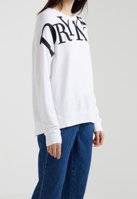 Vitt sweatshirt i mjukt tyg med stark svart text. Har rundad halsringning och långa ärmar, kombinerat med blå jeans.