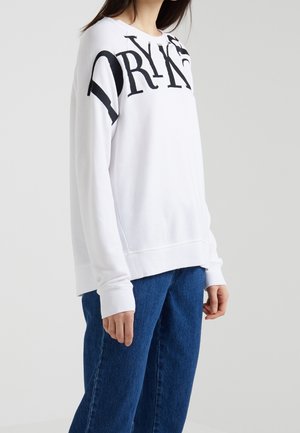 Vitt sweatshirt i mjukt tyg med stark svart text. Har rundad halsringning och långa ärmar, kombinerat med blå jeans.