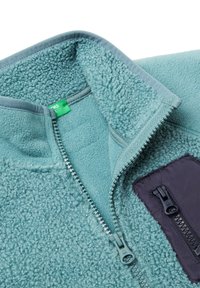 Giacca in fleece di un azzurro chiaro con una superficie texture. Presenta una chiusura con zip frontale e una tasca viola scuro con una zip coordinata.