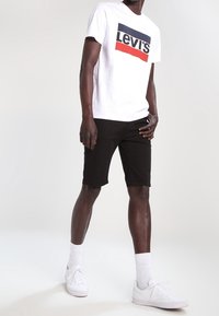 Vit bomull t-shirt med röd och blå "Levi's" logotyp, tillsammans med svarta shorts och vita sneakers. Enkel, avslappnad outfit med rena linjer.