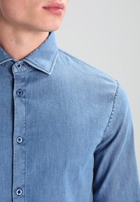 Ljusblå denimskjorta med krage som knäpps, med små blå knappar och en texturerad tyg, delvis synlig på modellens hals och axel.