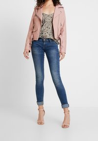 Giacca da moto in pelle rosa chiaro sopra un top con stampa leopardata, abbinata a jeans skinny blu e sandali con tacco beige. Design semplice e moderno.