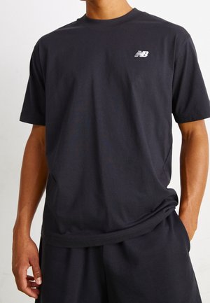 Person trägt ein einfaches schwarzes T-Shirt mit kurzen Ärmeln und kleinem weißen New Balance-Logo auf der Brust sowie passende schwarze Hose, Hände entspannt.