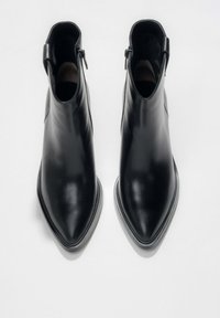 Para czarnych skórzanych botków za kostkę z szpiczastymi noskami i bocznymi zamkami, przedstawionych widokiem z góry na białym tle.