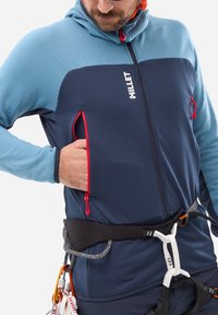 Technical climbing jacket, combining textured navy and light blue panels, features red zippered pockets, a fitted hood, and adjustable waistband.