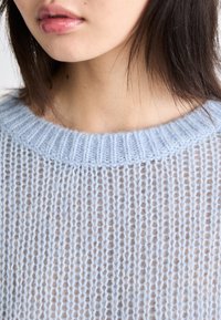 Pull en tricot bleu clair avec un large col côtelé et une texture ouverte et aérée, mettant en valeur un motif de tricot lâche.