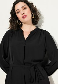 Vestido negro envolvente con manga larga, que presenta un escote en V y un cinturón anudado. Hecho de una tela suave, muestra una silueta relajada.