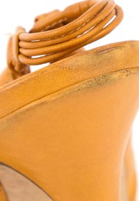 Gros plan d’une chaussure à talon haut en cuir marron clair usée, montrant des bords éraflés et une sangle de cheville nouée.