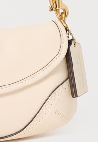 Cream leather handbag with a curved design, brown stitching accents, and a gold hardware clasp. Includes a detachable tag with branding.