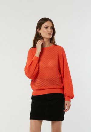 Pull en tricot orange vif avec un motif de losanges et un bord côtelé. Associé à une jupe noire, avec des manches longues légèrement bouffantes.